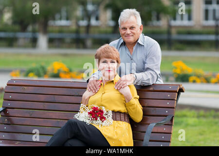 Ein älterer Mann sanft umarmt seine strahlende Frau auf einer Bank in einem Park. Stockfoto