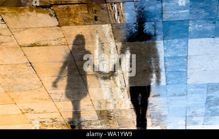 Unscharfe Reflexion Schatten Silhouetten von einem jungen Paar auf einer nassen Straße an einem sonnigen Sommertag in der Altstadt Stein Pflaster