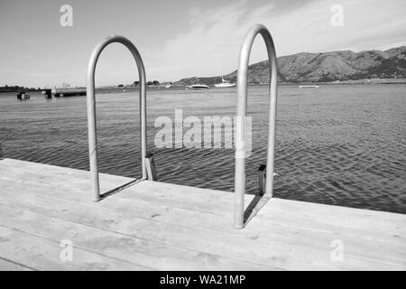 Ende eines hölzernen Pier mit einem metallischen Leiter nach unten, die in einem norwegischen Fjord mit Booten und Bergen im Hintergrund (schwarz und weiß) Stockfoto