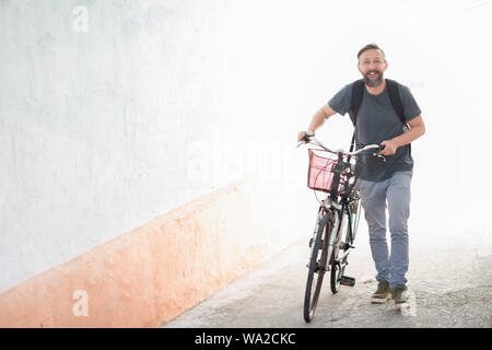 Eine junge lächelnde stilvolle bärtiger Hippie Mann mit einem Rucksack durch Drücken einer retro Fahrrad beim Gehen auf die Straße Stockfoto
