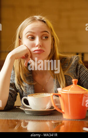 Junge Blondine mit einer Teekanne im Cafe sieht beiseite, sehnt sich vermisst, macht eine Grimasse Träume Stockfoto