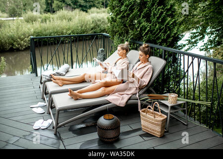 Zwei junge Frauen in der Bademäntel liegen auf den Liegen, entspannend und verbringt Zeit im luxuriösen Spa im Freien auf der Terrasse Stockfoto