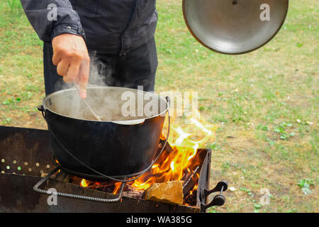 Küche bei Party Picknick geöffnet. Pilav Kochen am Feuer im Freien. Koch Koch ist das rühren Mahlzeit Teller in den großen Topf von Kelle. Bushpot bleibt auf Draht Rack und s Stockfoto