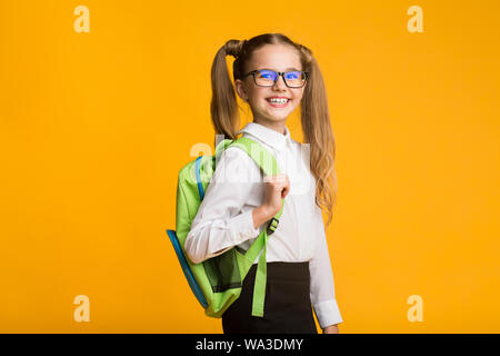 Lächelnd kleines Schulmädchen posiert mit Rucksack auf gelbem Hintergrund Stockfoto