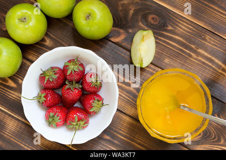 Urlaub der Sommer Ernte Hintergrund. Rote Erdbeeren in Weiß Schüssel und ganze Äpfel auf dunklen Holztisch mit kopieren. Gelber Honig im Glas Schüssel Stockfoto