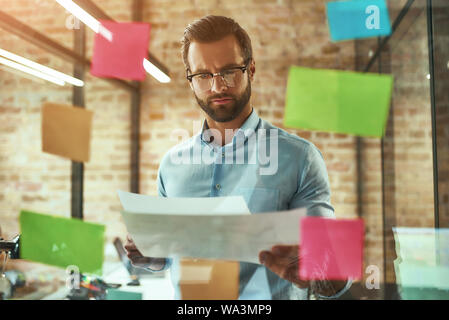 Kreative Ideen. Jung aussehenden bärtigen Mann in Brillen Planung und mit bunten Aufkleber beim Stehen vor der Glaswand. Projekt Management Stockfoto