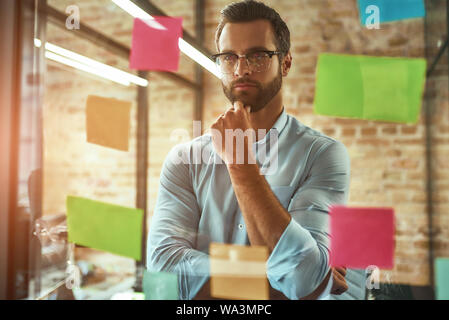 Nachdenken über Business. Jung aussehenden bärtigen Mann in Brillen Planung und mit bunten Aufkleber beim Stehen vor der Glaswand. Projekt Management Stockfoto