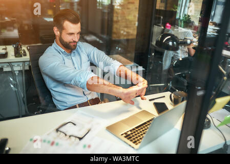 Entspannung am Arbeitsplatz. Müde junge Bartgeier Geschäftsmann in formale Abnutzung streckte seine Arme und lächelnd, während im modernen Büro zu sitzen. Arbeitsplatz. Pause Stockfoto