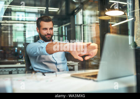 Kein Stress. Junge Bartgeier Geschäftsmann in formale Abnutzung streckte seine Arme und lächelnd, während im modernen Büro zu sitzen. Arbeitsplatz. Pause Stockfoto