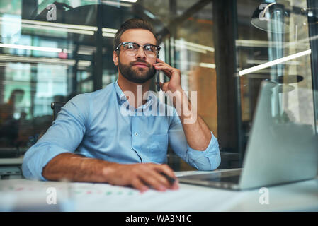 Gerne helfen. Porträt der jungen gutaussehenden Mann in Brillen und formale Abnutzung sprechen am Telefon mit Kunden, während im Büro zu sitzen. Arbeitsplatz. Business Konzept Stockfoto