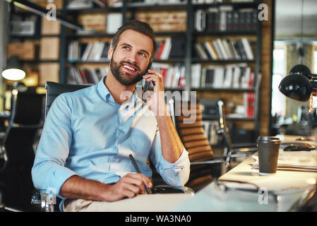 Glücklich zu hören, dass Sie junge Bartgeier Geschäftsmann in formale Abnutzung im Gespräch mit dem Kunden per Telefon und lächelnd während der Sitzung in moderne Büro. Arbeitsplatz. Business Konzept Stockfoto