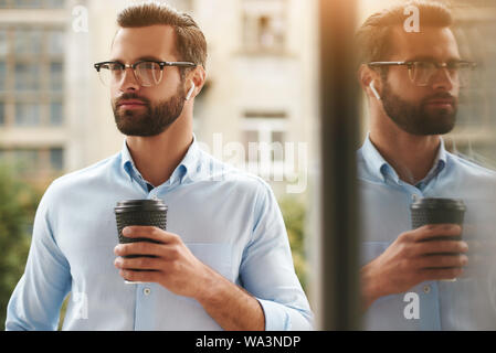 Porträt der jungen und hübschen bärtiger Mann mit Brille und Kopfhörer holding Tasse Kaffee und Wegsehen im Stehen im Büro Balkon Stockfoto