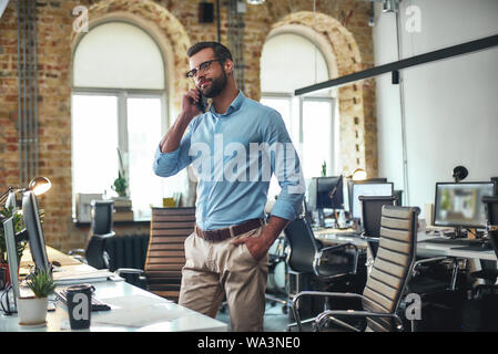 Gute Nachrichten. Porträt der jungen und fröhlichen bärtigen Mann in Brillen mit smart phone mit Client beim Stehen im modernen Büro. Kommunikation. Business Konzept Stockfoto