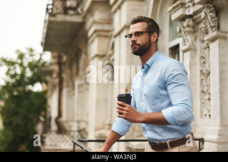 Jung aussehenden bärtigen Mann in Brillen und Gesellschaftsmode holding Tasse Kaffee und weg schauen, während Sie im Büro Balkon stehen. Kaffeepause Stockfoto