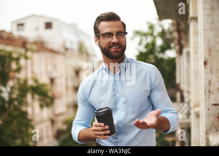 Jung aussehenden bärtigen Mann mit Brille und Kopfhörer holding Tasse Kaffee und Gespräch mit Freund, während Sie im Büro Balkon stehend Stockfoto