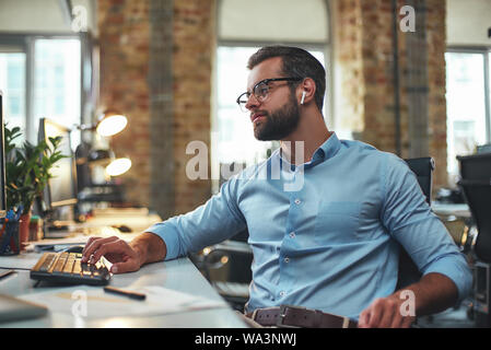 Anstrengenden Tag. Seitenansicht des Jungen bärtigen Mann mit Brille und Kopfhörer etwas Tippen auf Computer, während im modernen Büro zu sitzen. Arbeit Konzept. Business Konzept Stockfoto