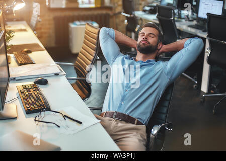 Zeit sich zu entspannen. Junge Bartgeier Geschäftsmann zurück lehnte sich mit den Händen hinter dem Kopf und entspannend, während im modernen Büro sitzen zufrieden. Ruht. Arbeitsplatz Stockfoto