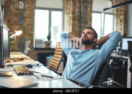 Genießen Sie brechen. Junge Bartgeier Geschäftsmann zurück lehnte sich mit den Händen hinter dem Kopf und entspannend, während im modernen Büro sitzen zufrieden. Entspannen Konzept. Arbeitsplatz Stockfoto