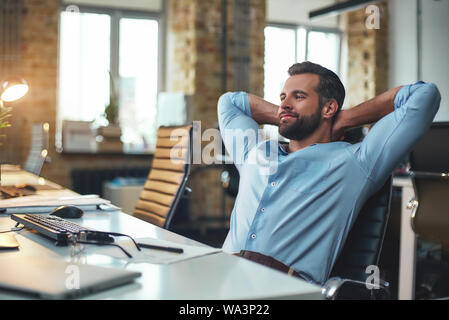 Arbeit geleistet. Junge Bartgeier Geschäftsmann zurück lehnte sich mit den Händen hinter dem Kopf und entspannend, während im modernen Büro sitzen zufrieden. Entspannen Konzept. Arbeitsplatz Stockfoto