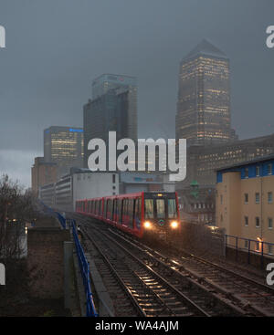 Westferry, London Docklands Light Railway 111 + 136 + 104 an trüben regnerischen Tag Ankunft in einem Regensturm mit einer Bank, Canary Wharf ist hinter Stockfoto