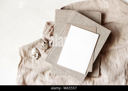 Feminine Schreibwaren, desktop Mock-up-Szene. Leere Grußkarte, Handwerk Umschlag, Muscheln und alte Bücher über beigefarbene Bettwäsche Tischdecke Hintergrund. Flach Stockfoto