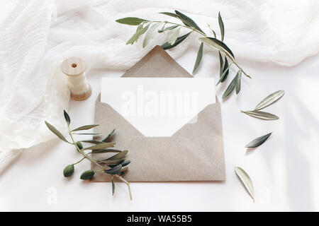 Feminine Hochzeit Schreibwaren, desktop Mock-up-Szene. Leere horizontale Grußkarte mit Handwerk Umschlag, Seidenband und Ölzweige. weißer Tisch Stockfoto