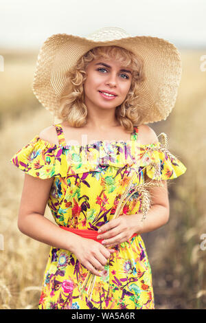 Porträt von einem schönen Mädchen in einen Hut holding Reif ährchen von Gerste in den Händen. Ein Modell in einer farbigen sundress Posen auf dem Hintergrund einer agric Stockfoto