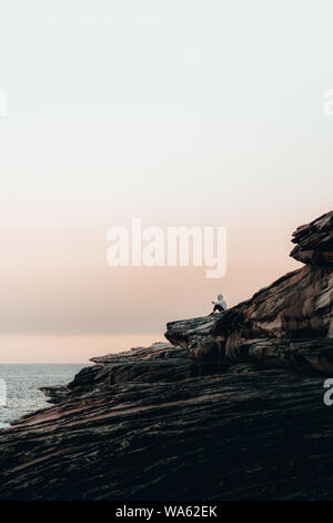 Mann in Hoodie sitzt auf dem Felsen, nachdenklich, während Sie den Sonnenuntergang am Trenerry finden, Coogee. Stockfoto