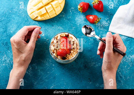 Draufsicht auf die Hände der Mädchen mit Löffel essen Dessert mit Mango, Müsli, Quark Mousse und Erdbeeren auf blauem Hintergrund Konzept der gesunde Desserts Stockfoto