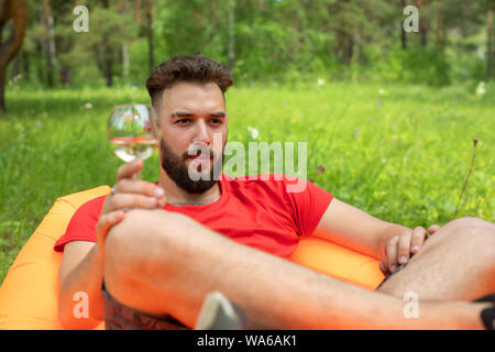 Freizeitaktivitäten Konzept, Camping, Sommerferien. Junger bärtiger Mann der kaukasischen Aussehen mit Gläsern Wein zur Festlegung von Orange Sofa. Entspannen und Genießen Stockfoto