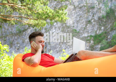 Student, bärtiger junger Mann sitzt auf einem aufblasbaren Sofa. Sieht auf einem Laptop, einem Lächeln und Gespräche am Mobiltelefon. hellen Sommertag Stockfoto