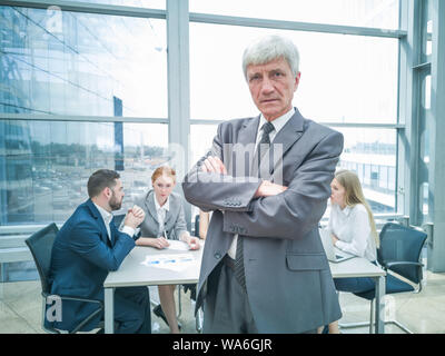 Zuversichtlich reife Geschäftsmann stand mit verschränkten Armen lächelnd, Team von seinen Kollegen an dem Tisch im Hintergrund sitzen Stockfoto