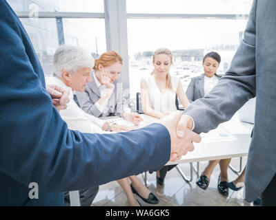 Handshake von Geschäftspartnern nach Diskussion der Finanzberichte, ihr Team im Hintergrund Stockfoto
