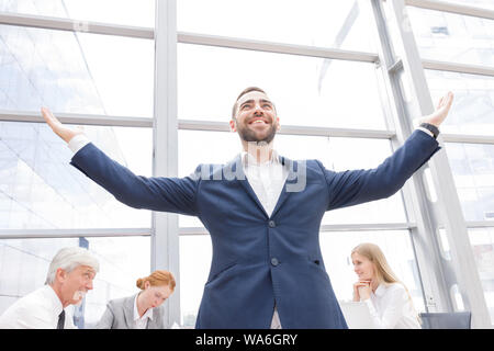 Junge freudige Geschäftsmann stehend mit erhobenen Armen lächelnd, Team von seinen Kollegen an dem Tisch im Hintergrund sitzen Stockfoto
