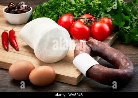 Türkische bryndza Schafskäse auf Holztisch mit Holz Schneidebrett mit fermentierten Würstchen, Eier, Olivenöl, Petersilie und Tomaten. Stockfoto