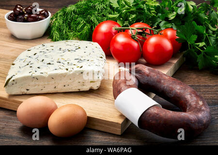 Türkisch Van Milch Käse mit Kräutern auf Holztisch mit Holz Schneidebrett mit fermentierten Würstchen, Eier, Olivenöl, Petersilie und Tomaten. Stockfoto