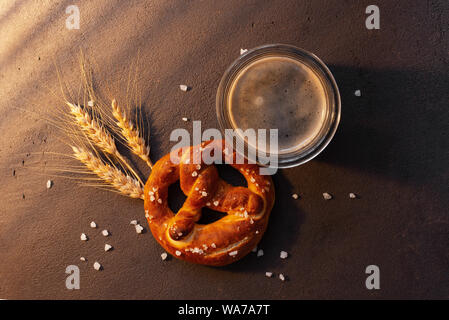 Gesalzene Brezel und Bierkrug auf dem Braunen strukturierte Tabelle mit Weizen Ohren eingerichtet, warme Sonne Licht Stockfoto