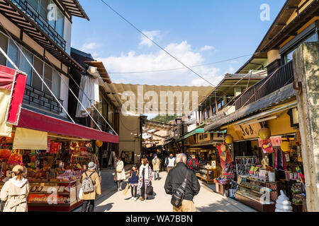 Japan, Miyajima. Blick entlang der Hauptstraße mit typischen touristischen Geschäften und Läden auf beiden Seiten, mit über Sonnenschutz gezogen, und Menschen zu Fuß. Stockfoto