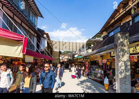 Japan, Miyajima. Blick entlang der Hauptstraße mit typischen touristischen Geschäften und Läden auf beiden Seiten, mit über Sonnenschutz gezogen, und Menschen zu Fuß. Stockfoto