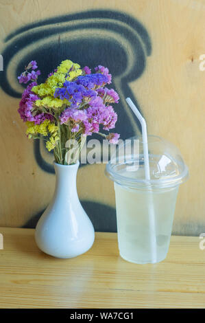 Limonade und Blumen. Limonade in einer Kunststoff Glas mit einem Deckel und einem Strohhalm. Im Sommer trinken. Wildblumen in einer Vase auf einem Holztisch. Bäckerei und qu Stockfoto