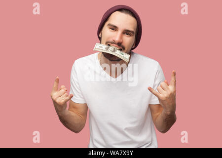 Crazy zuversichtlich aussehenden bärtigen Jungen hipster Mann im weißen Hemd und casual Hut, die hielten viele Dollar auf die Zähne, die Rocker unterzeichnen. Indoor, Stockfoto