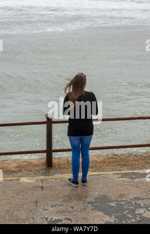 Eine junge Frau mit langen Haaren neben Geländer am Meer Blick Meer an einem stürmischen rauhen Tag am Meer. Stockfoto