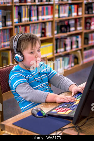 Ein kleiner Junge mit Kopfhörern, spielt auf einer Lap Top an der lokalen öffentlichen Bibliothek Stockfoto