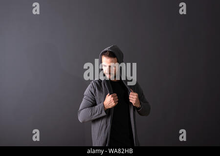 Eine ernsthafte hübscher junger Mann in einem grauen Kapuzenpullover vor einem dunkelgrauen Hintergrund in einem Studio. Stockfoto