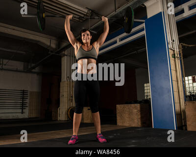 Eine sportliche kurze behaarte Frau mit starken abs tut Gewichtheben mit einem barbell in ein Fitnessstudio. Die muskulöse Frau tut eine Schulter, an der Oberleitung reinigen. Stockfoto