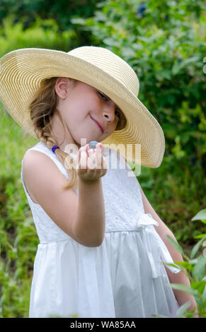 Eine liebenswerte Kind in einer Diskette Strohhut ist heraus halten, frische Blaubeeren zu Probe Stockfoto