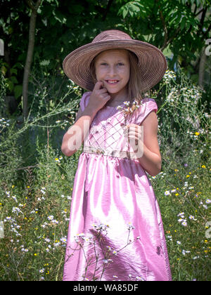 Ein junges Mädchen schüchtern stellt in einem Feld von Gänseblümchen, trägt einen Schlapphut und rosa Kleid Stockfoto