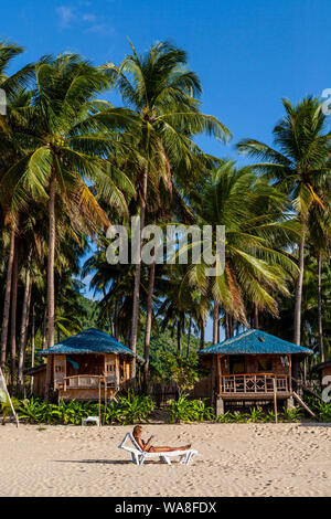 Eine weibliche Touristen auf einer Sonnenbank ein Buch lesen außerhalb Ihrer Unterkünfte direkt am Strand, Nacpan Beach, El Nido, Palawan, Philippinen. Stockfoto