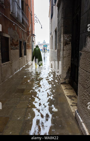 Ein caped Tourist, an einem regnerischen Tag, zu Fuß durch eine überflutete Straße während der Acqua Alta (hohe Wasser) Fall, Calle de la Rasse, Venedig, Italien Stockfoto