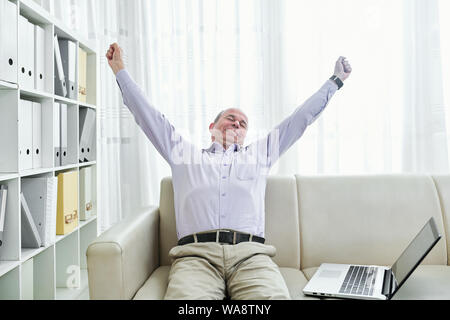 Müde, aber glücklich im mittleren Alter Geschäftsmann Sitzung vom Sofa und Stretching nach Arbeiten am Laptop den ganzen Tag lang Stockfoto